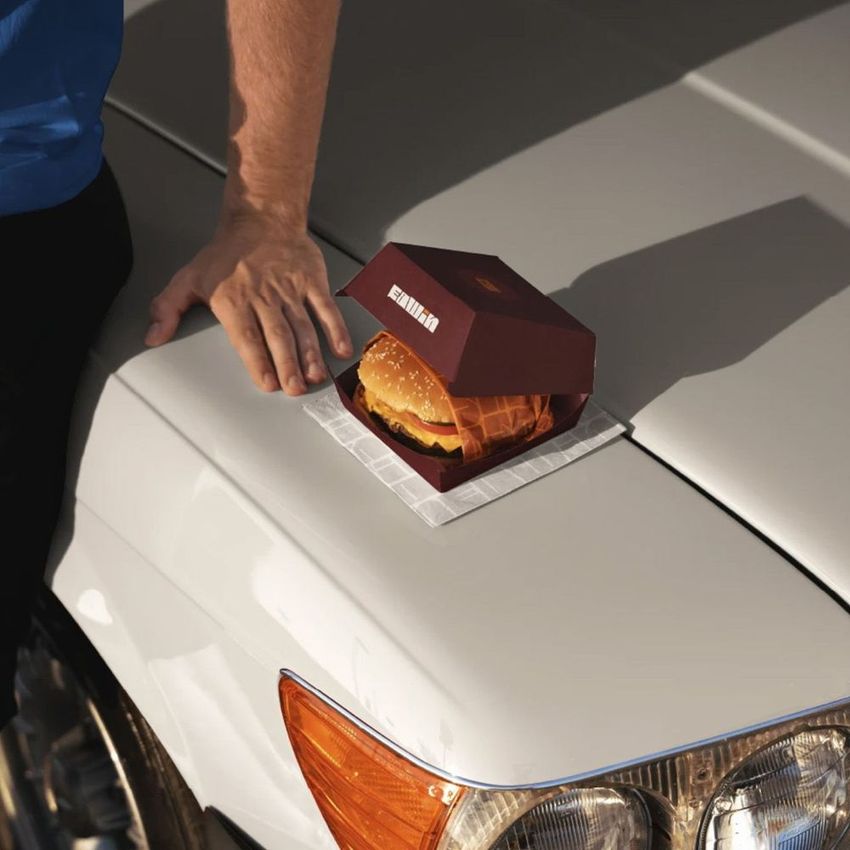 Burger box mockup featuring a burger in a maroon container placed on the hood of a white car, with a person's arm nearby.