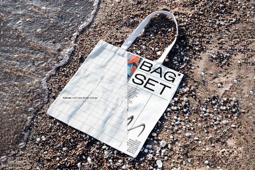 Canvas tote bag mockup on a shoreline, showing a split design with grid pattern and bold text, top-down perspective.
