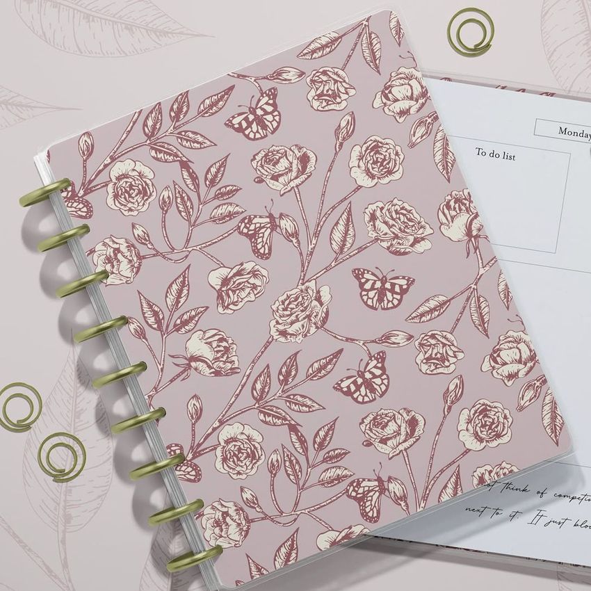 Top view of two spiral planners on a light surface, one closed with a pink floral and butterfly cover, the other open showing lined pages.