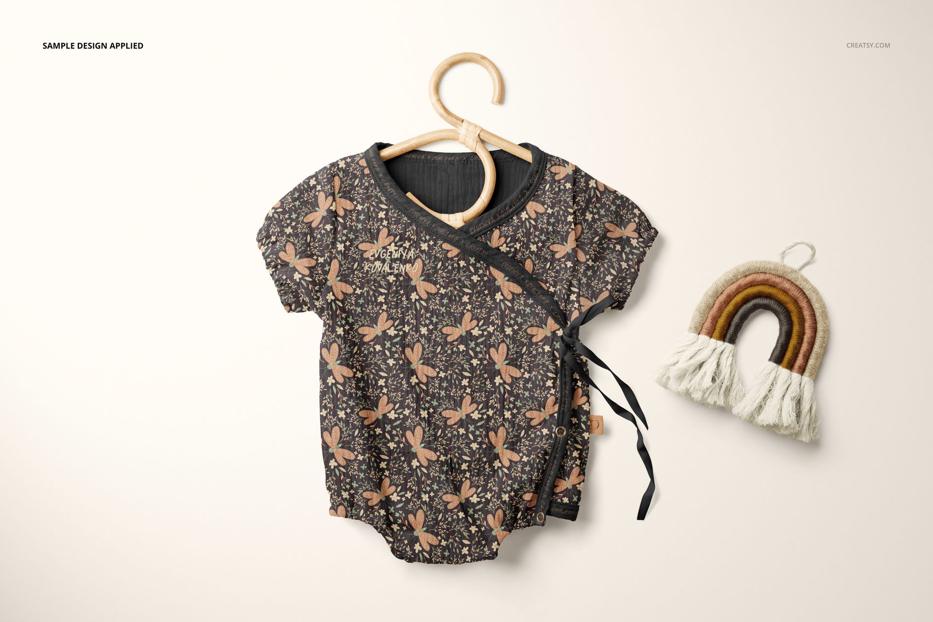 Front view of a short-sleeve muslin onesie mockup with a floral pattern on a wooden hanger, next to a rainbow-shaped decor.