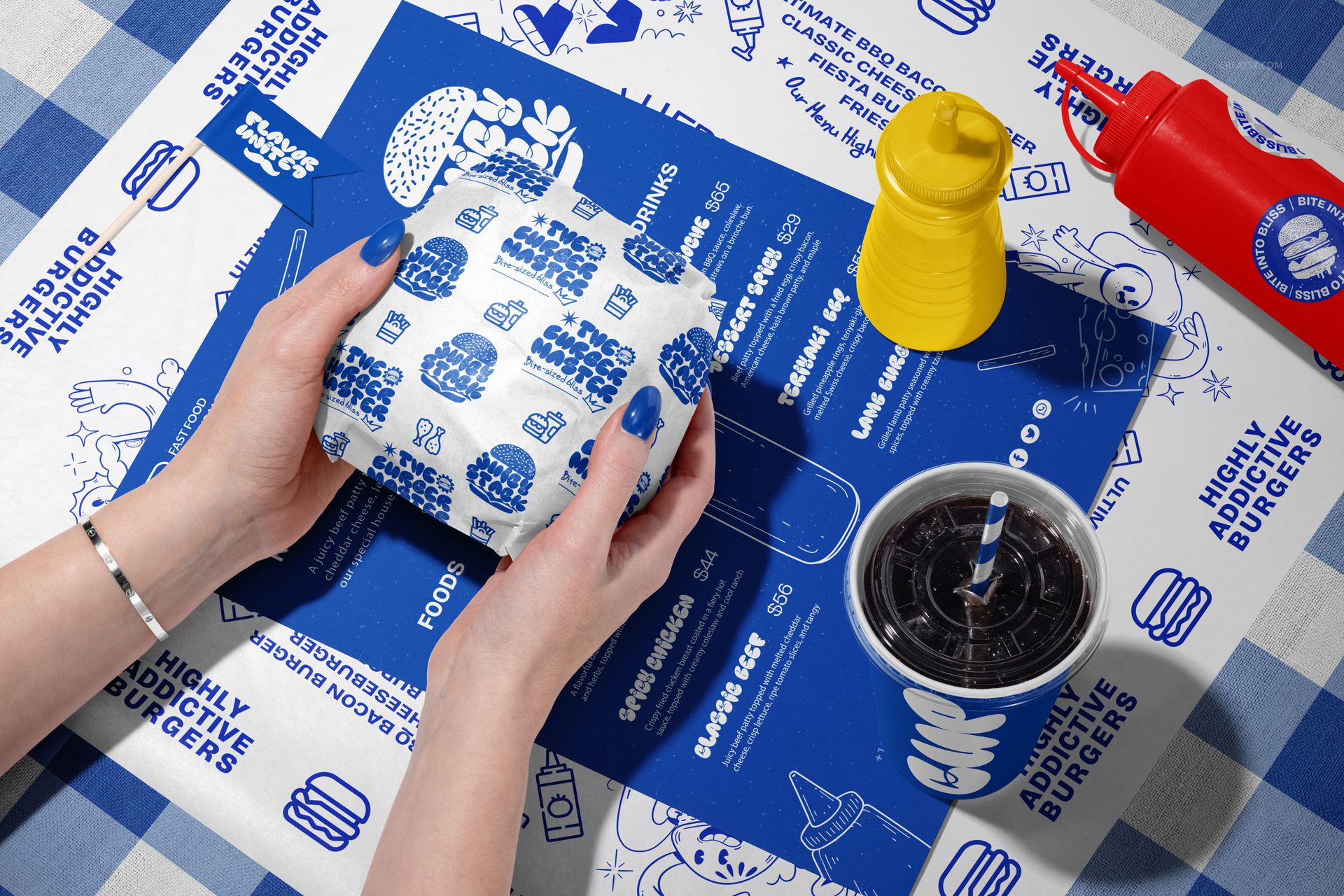 Hands holding a wrapped vegan burger over blue and white branded packaging mockups, with drink cup and condiments on a checkered tablecloth.