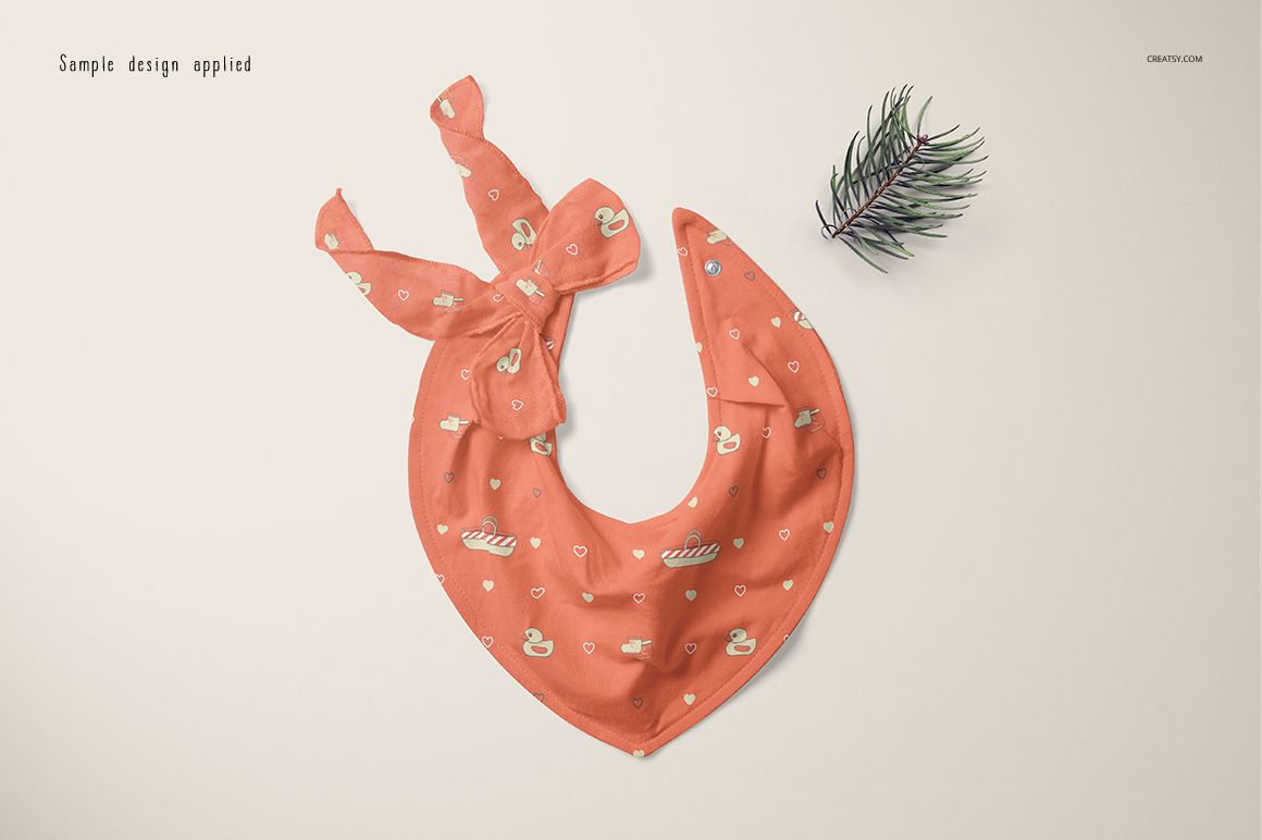 Coral baby bandana bib mockup featuring swan and heart motifs, shown from above with a decorative branch beside it.