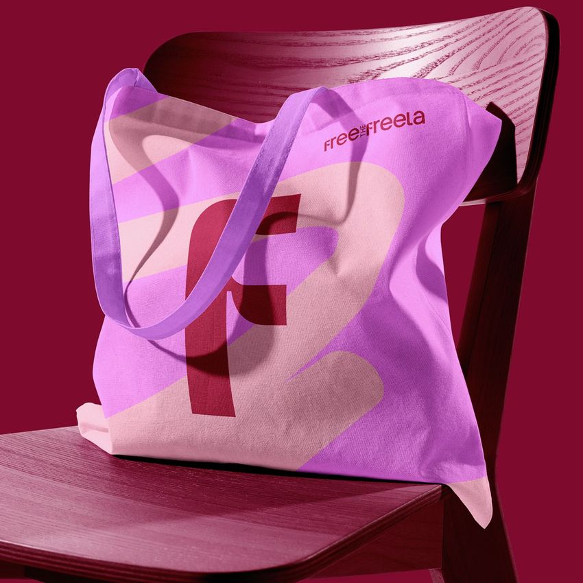 Tote bag mockup featuring a pink and purple design with bold abstract shapes, draped over a wooden chair against a burgundy background.