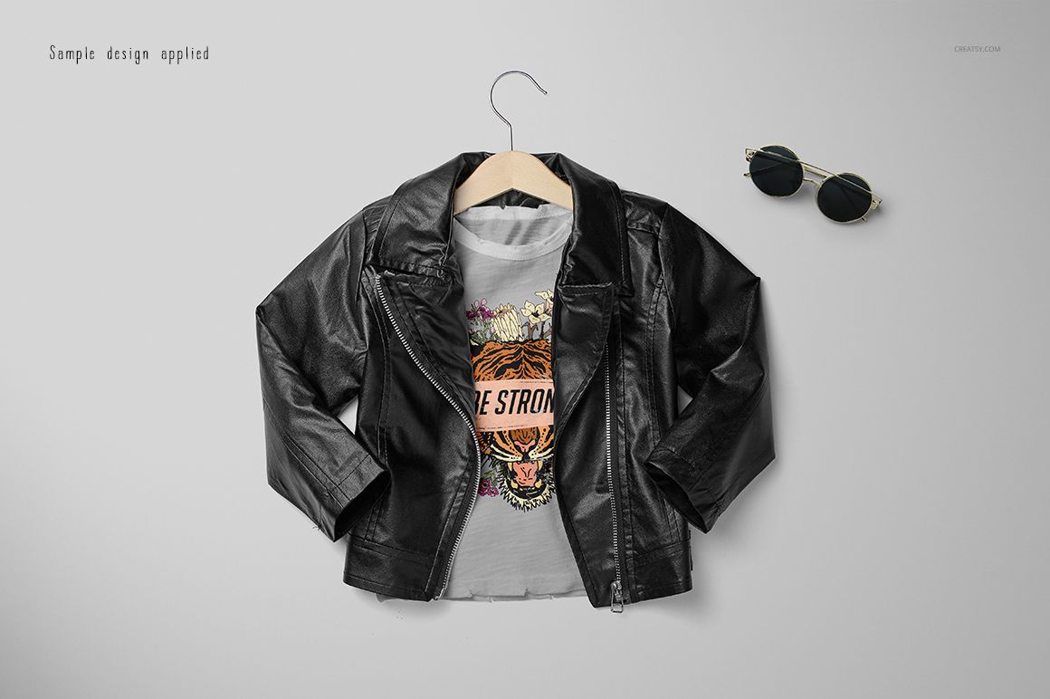 Front view of a girls chic set mockup featuring a black faux leather jacket on a hanger over a graphic t-shirt, with sunglasses nearby.