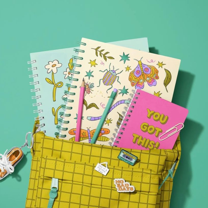 School backpack mockup with spiral notebooks featuring colorful bug and flower illustrations, pens, and a small sneaker keychain.