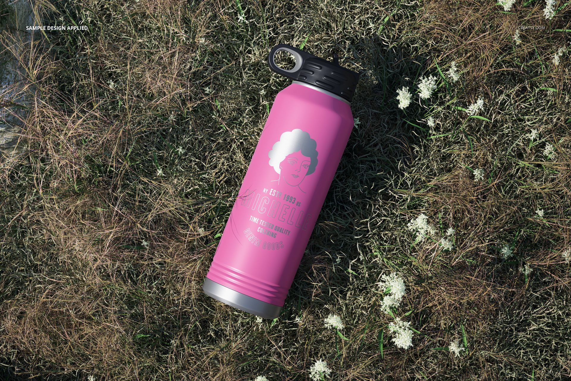Powder coated pink water bottle mockup with black handle cap placed on grassy ground with scattered white blossoms.