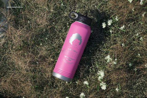 Powder coated pink water bottle mockup with black handle cap placed on grassy ground with scattered white blossoms.