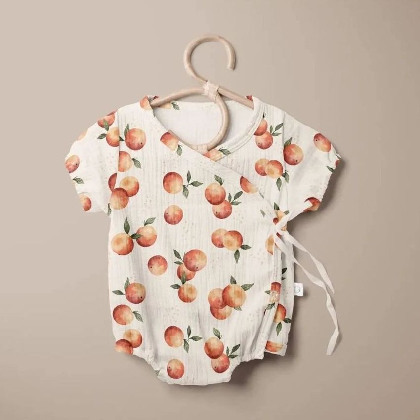 Muslin onesie mockup featuring a short-sleeved baby bodysuit with an orange fruit pattern, displayed on a wooden hanger against a beige background.