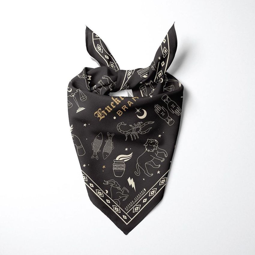 Pet bandana mockup featuring a black fabric with gold zodiac illustrations and geometric border, displayed on a white background.