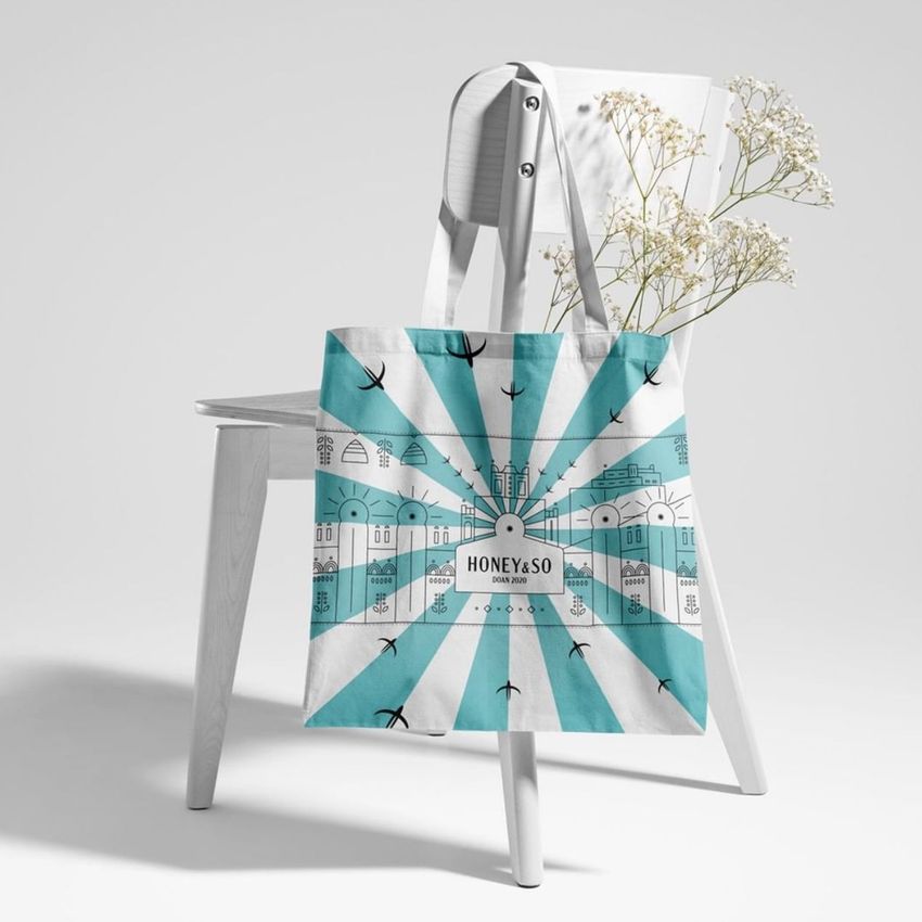 Tote bag mockup displayed on a white chair, featuring a blue and white geometric cityscape design and a text overlay in the center.
