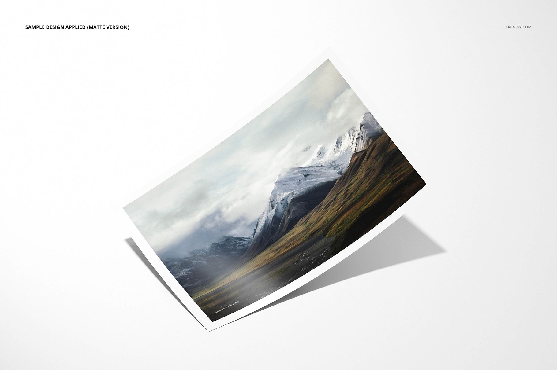 Matte photo print mockup featuring a landscape image with mountains and grassy fields, angled on a white surface.