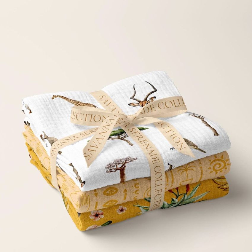 Stack of two crinkle muslin swaddle blankets, one white with safari animal prints and one yellow with botanical patterns, tied with a ribbon.
