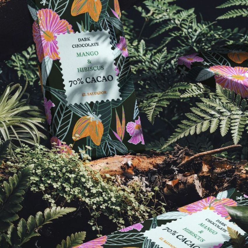Chocolate box mockup featuring tropical floral print packaging, placed among lush green ferns and moss in a forest setting.