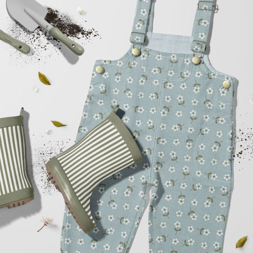 Kids dungarees mockup with a light blue floral pattern, striped green rain boots, and gardening tools on a white surface.