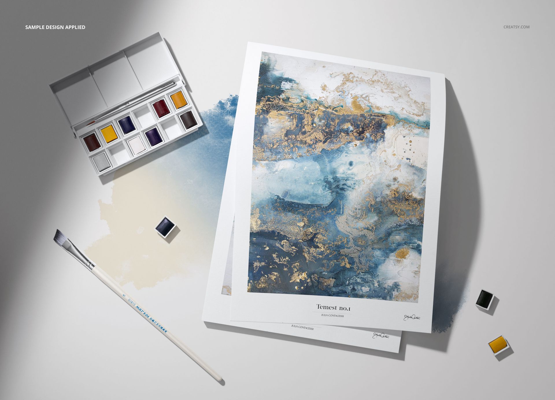 Top view of a watercolor paint set, brush, loose paint pans, and mockup paper with a blue and gold abstract design.