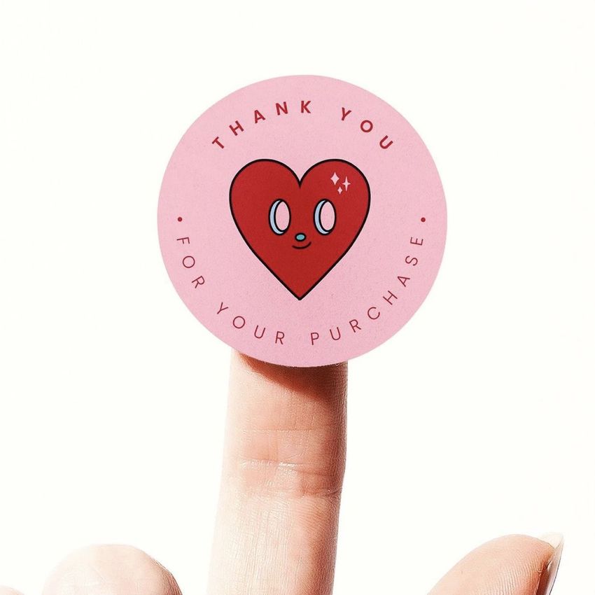 Round pink sticker mockup with a red heart illustration and text, displayed on the tip of an upright finger against a white background.