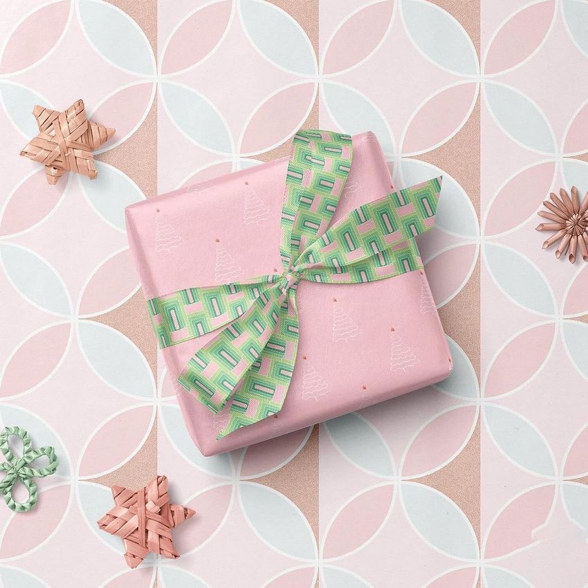 Square gift box mockup wrapped in pink paper with white tree patterns, tied with a green geometric ribbon, on a pastel geometric background.
