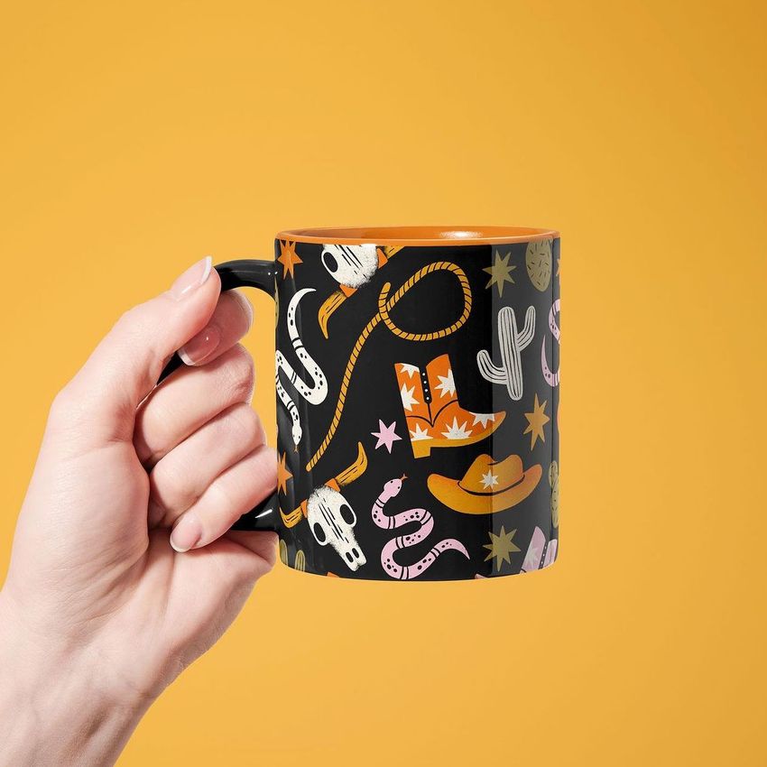 Hand holding a mug mockup with a cowboy-themed pattern featuring boots, hats, cacti, and stars on a black background.
