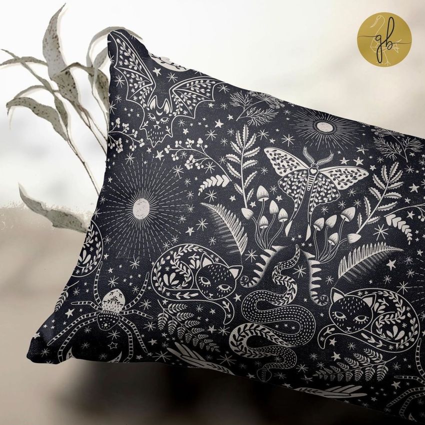 Rectangular pillow mockup featuring a dark fabric with white mystical illustrations of bats, snakes, and stars, set on a neutral surface with a leafy plant in the background.