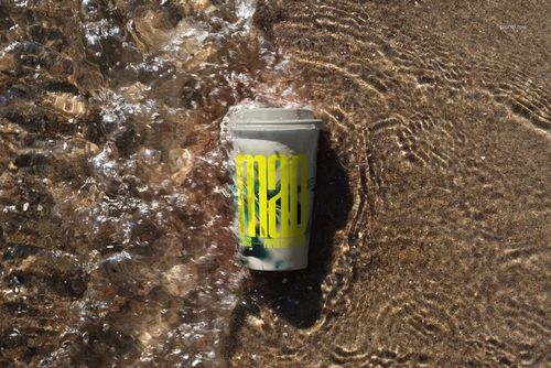 Mockup of a paper coffee cup with a white lid on sandy beach, water washing over the cup from the left side.