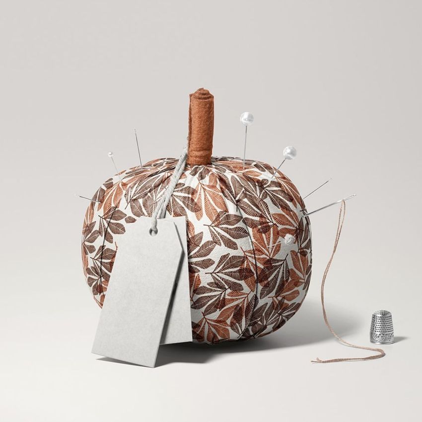 Fabric pumpkin mockup with brown and white leaf pattern, sewing pins, blank tag, and thimble on a light background.