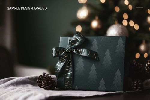 Christmas gift box mockup with satin ribbon, dark green and silver tree design, placed beside pinecones and blanket.
