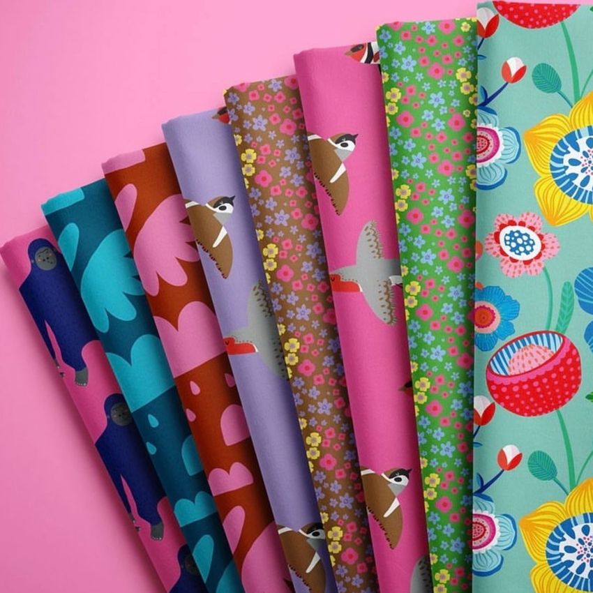 Folded fabrics mockup featuring seven pieces of fabric with colorful floral and bird patterns arranged on a pink background.