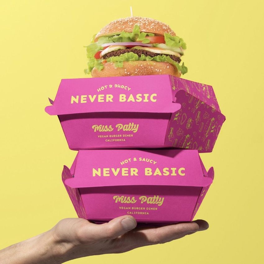 Packaging mockup featuring two stacked pink burger boxes with bold text, held in a hand against a yellow background and topped with a burger.