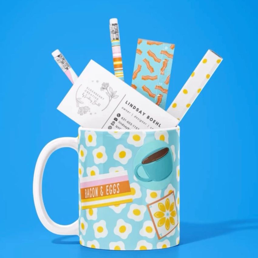 Mug mockup with a playful bacon and eggs design, filled with pencils, business cards, and stationery on a blue background.