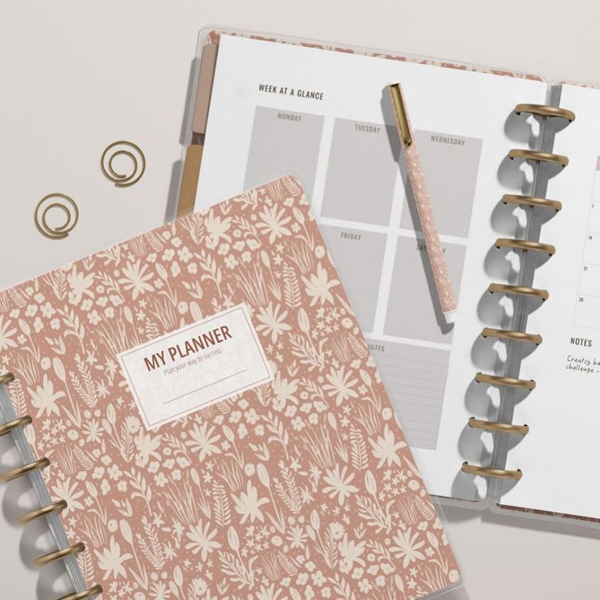 Top view of two spiral planners with pink floral covers, open to weekly pages, with a pen and gold paper clips nearby.