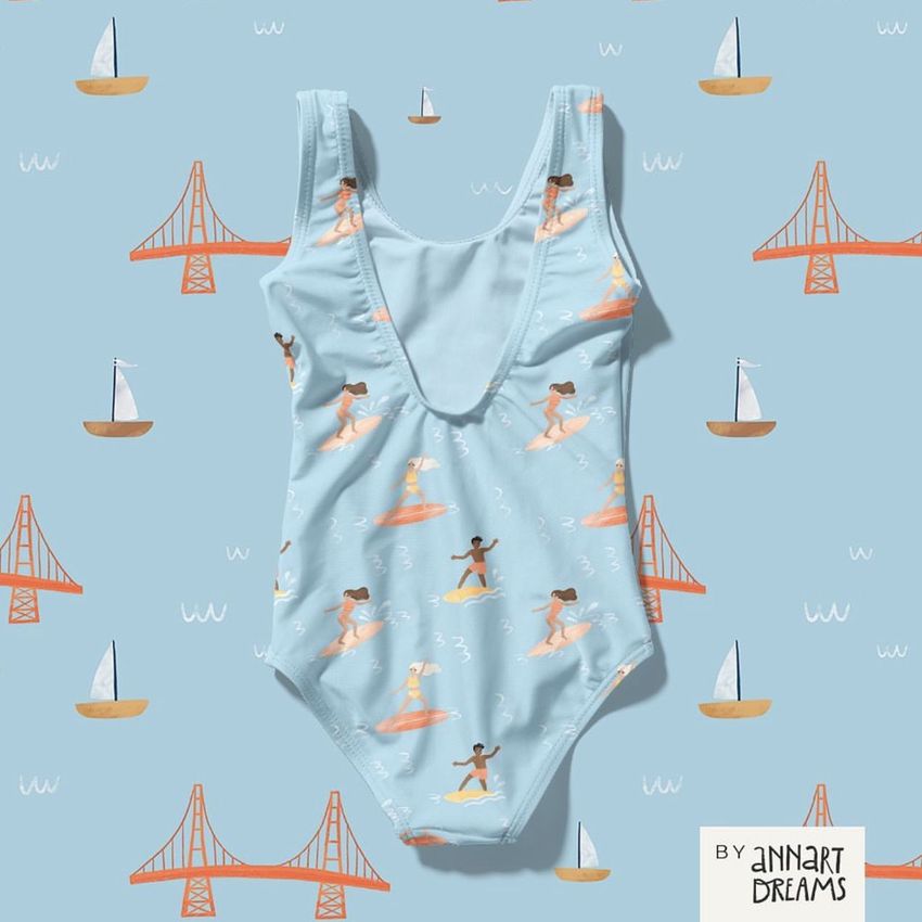 Baby swimsuit mockup displayed on a blue background with sailboat and bridge illustrations, featuring a surfing pattern.