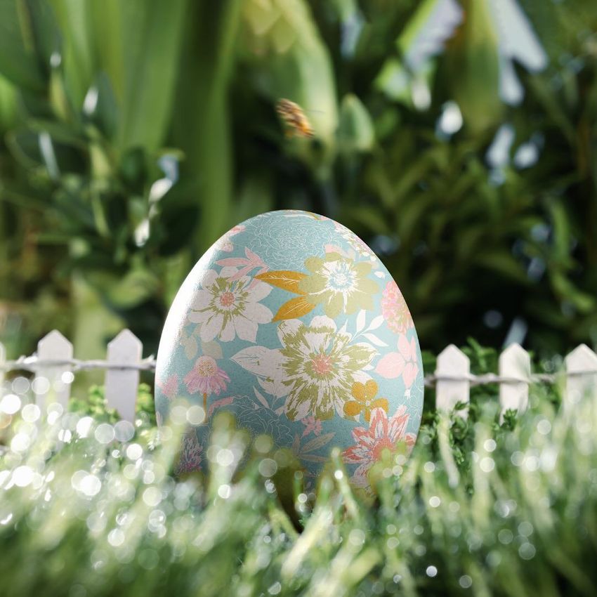 Easter egg card mockup featuring a pastel egg with floral patterns standing in grass, with a white picket fence and greenery in the background.