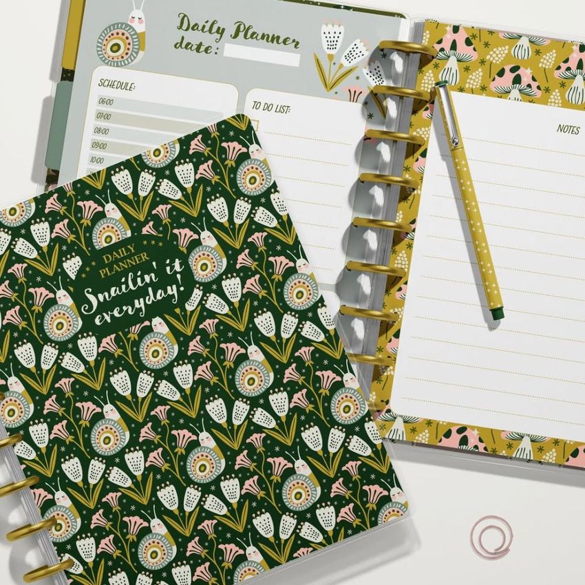 Top view of two spiral planners with floral covers, one open to a daily planner page and one closed, on a white surface.