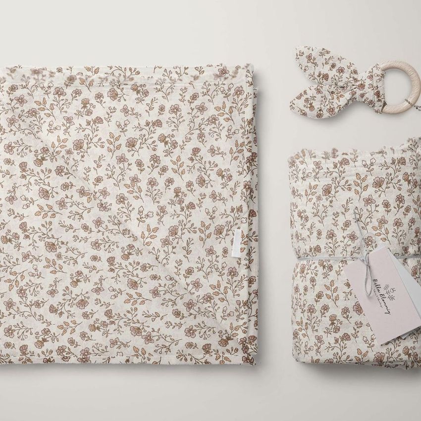 Muslin swaddle blanket mockup featuring a delicate floral pattern in soft brown and beige tones, shown folded and rolled with a wooden teether on a light background.