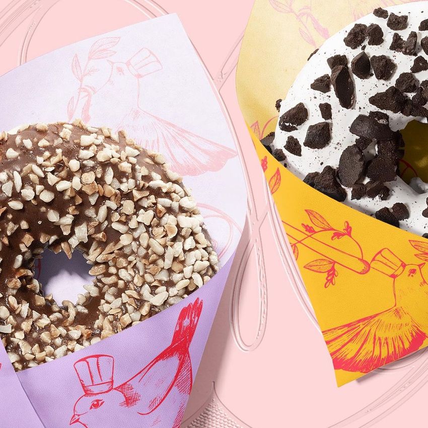 Food safe paper mockup featuring two donuts wrapped in pastel and yellow printed sheets with bird illustrations on a pink background.