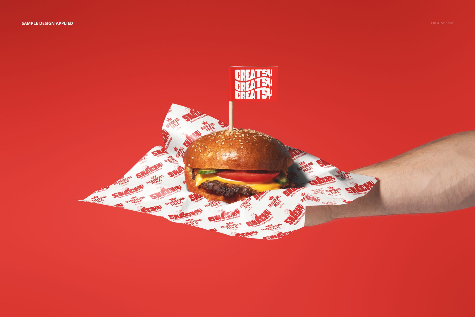 PSD mockup showing a burger on branded wrapping paper, presented on an outstretched hand against a red backdrop.