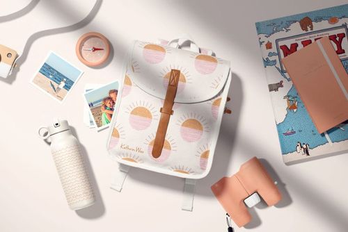 Kids backpack mockup with sunrise pattern, surrounded by travel props like water bottle, compass, and postcards on a light surface.