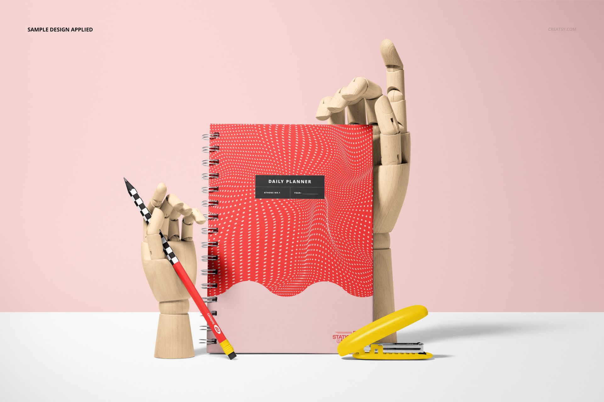 Stationery mockup featuring a spiral planner with a bold red design, wooden mannequin hands, and a yellow stapler.