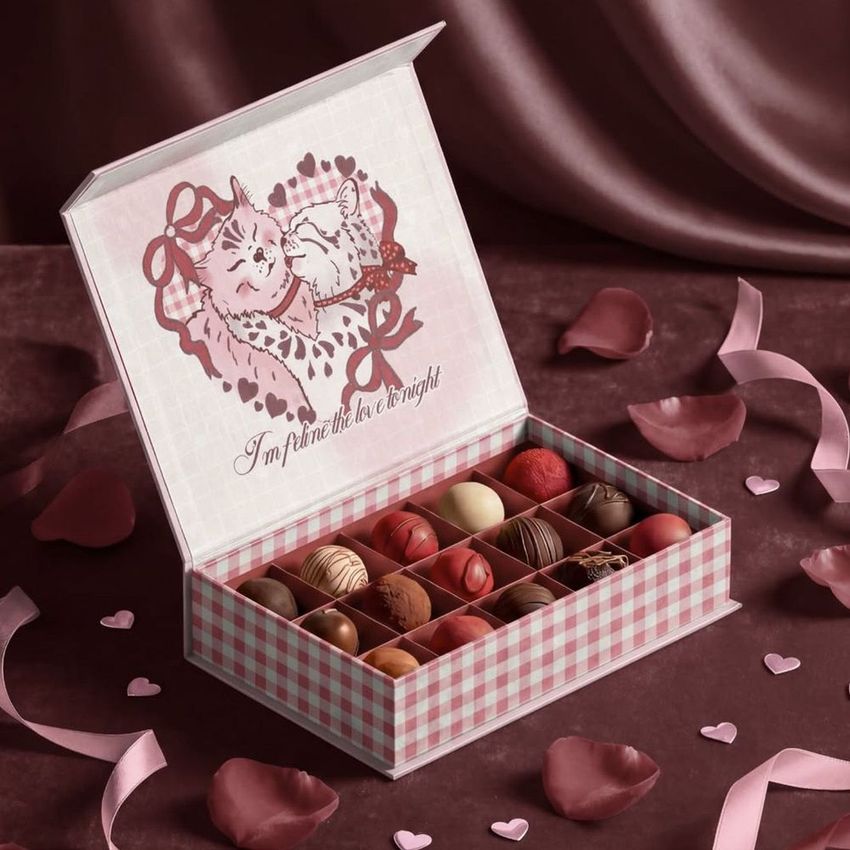 Open chocolate box mockup with assorted pralines arranged in a grid, set on a pink fabric surface with scattered rose petals.