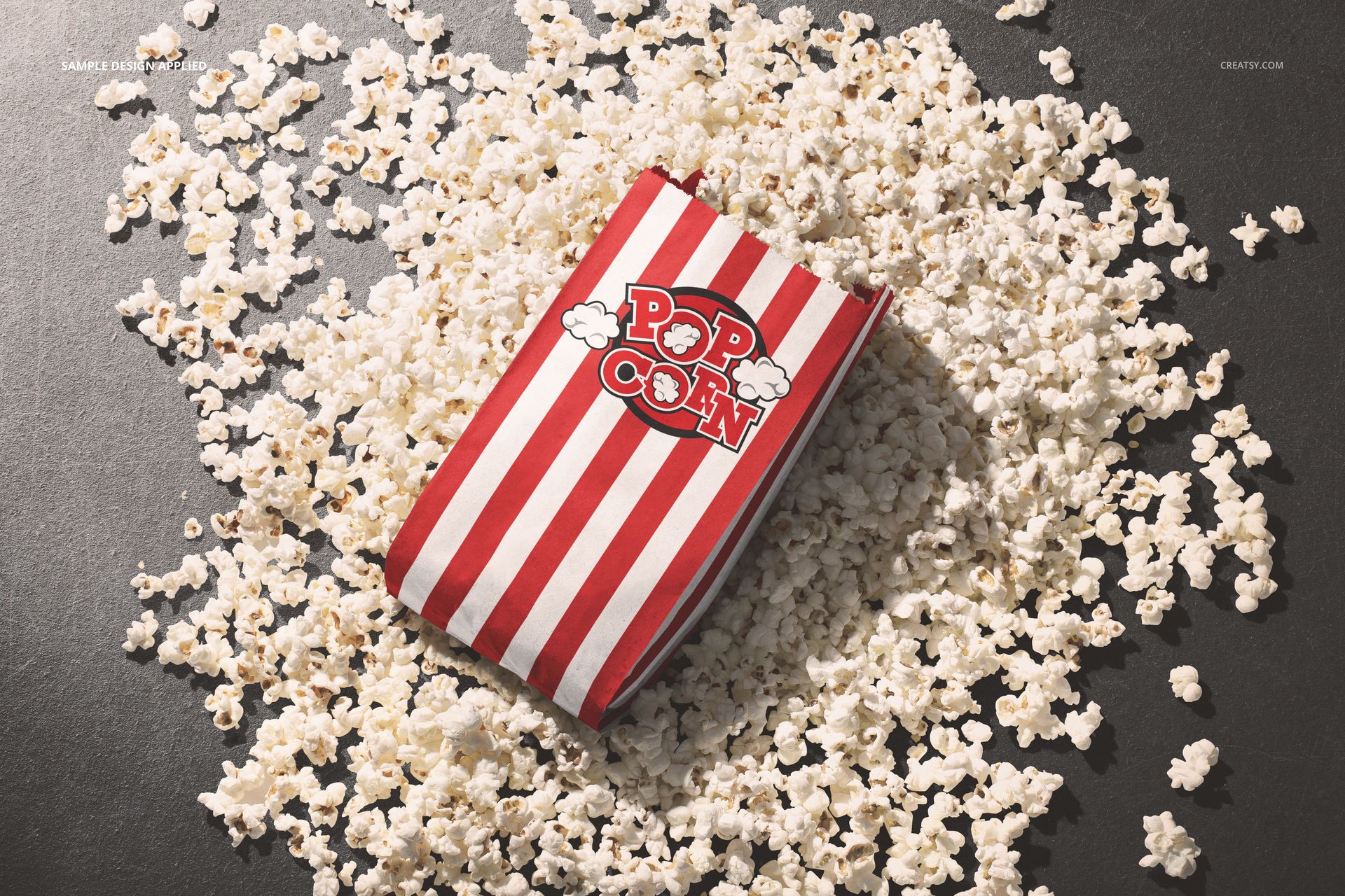 Red and white striped popcorn paper bag mockup lying on a dark surface surrounded by scattered popcorn pieces.
