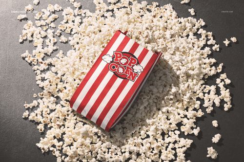 Red and white striped popcorn paper bag mockup lying on a dark surface surrounded by scattered popcorn pieces.