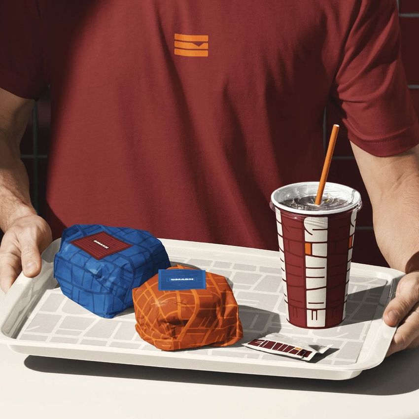 Man holding a tray mockup with two wrapped burgers, a cup with a straw, and a condiment packet on a tiled surface.