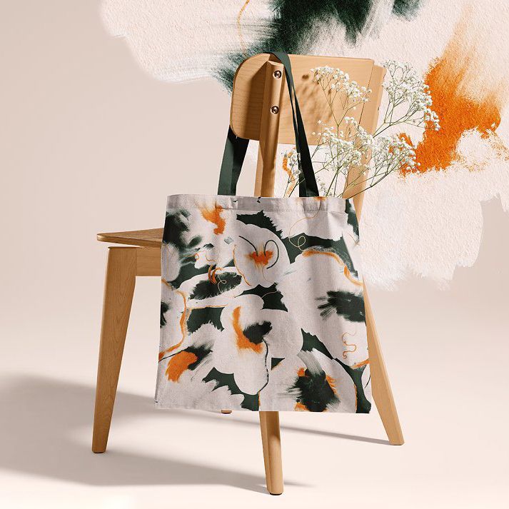 Tote bag mockup featuring a fabric bag with abstract floral patterns in green, orange, and white, hanging on a wooden chair.