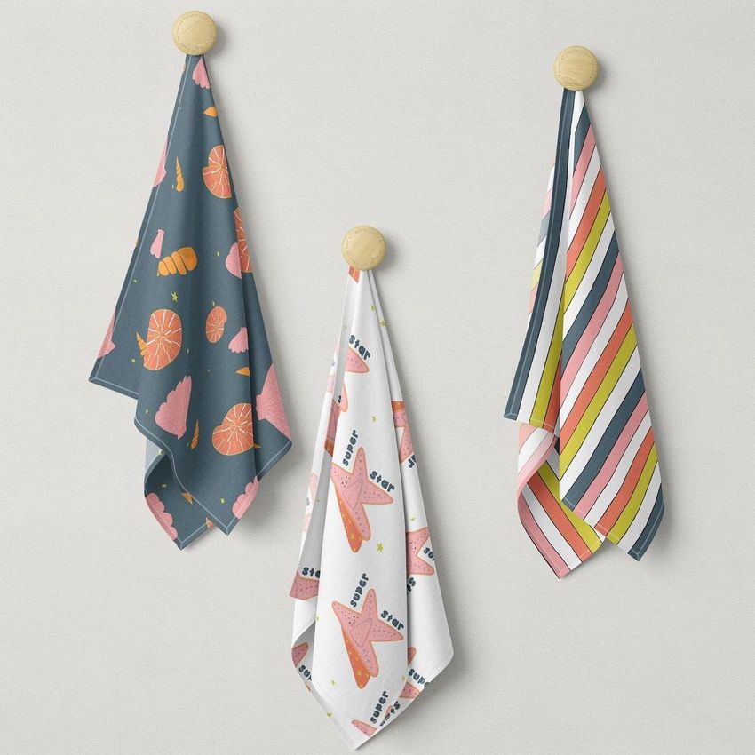 Three hanging fabric mockups displayed on wooden hooks, featuring colorful patterns and a neutral wall background.