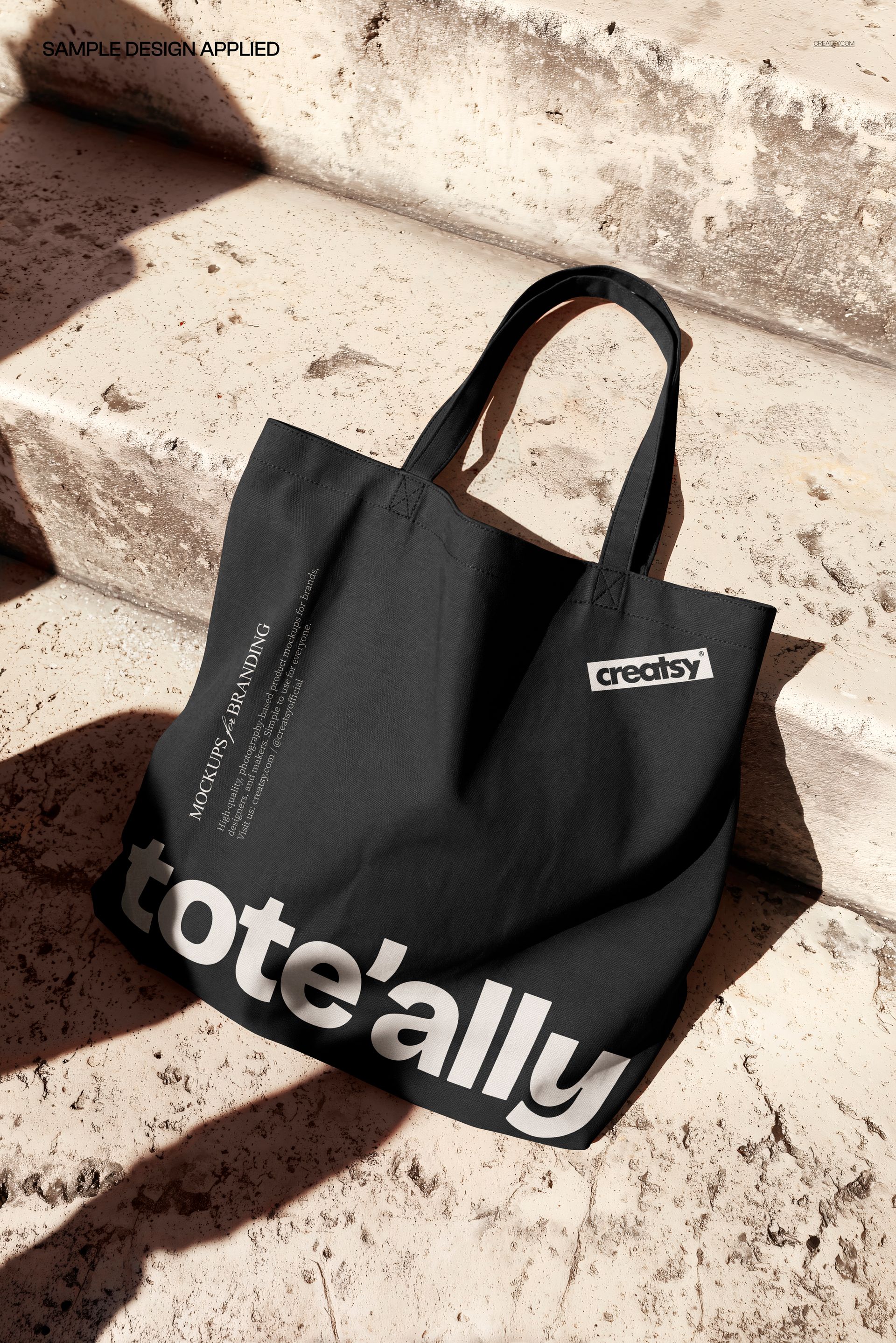 Canvas tote bag mockup in black with large white lettering, displayed on textured stone stairs in natural light.