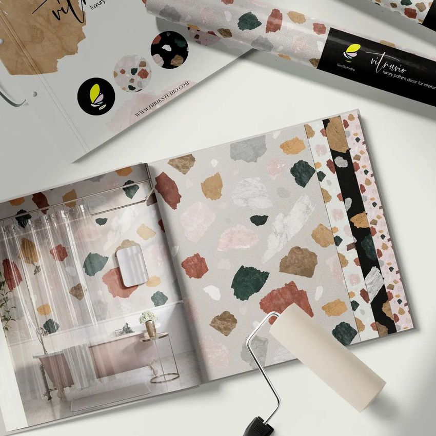 Wallpaper sample book mockup showing an open page with terrazzo patterns, a paint roller, and rolled wallpaper on a white surface.