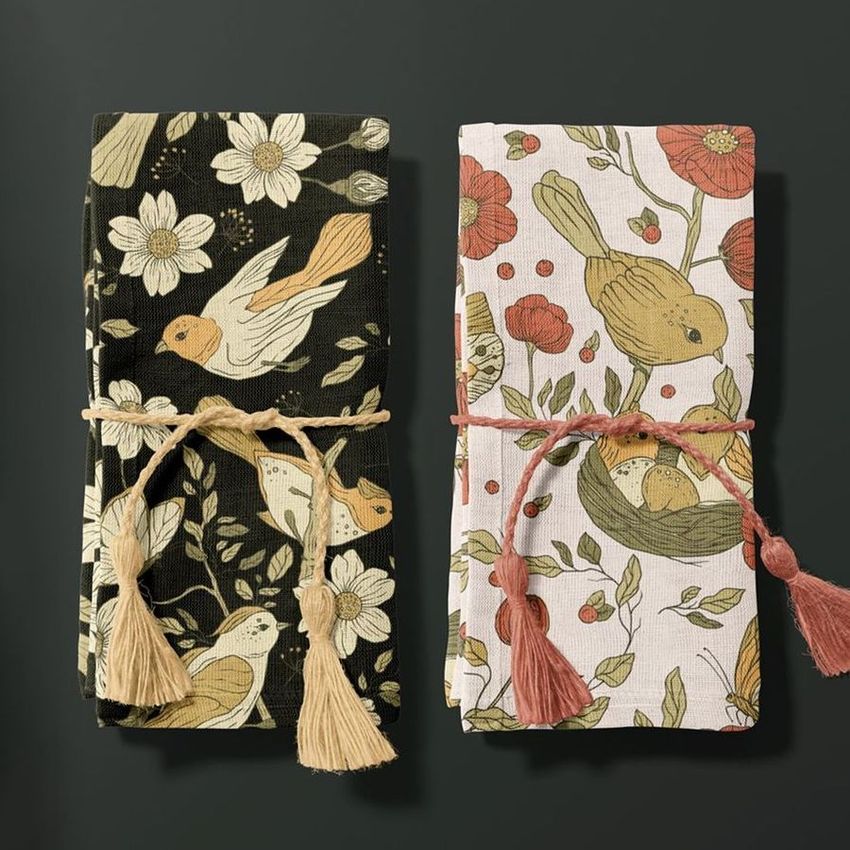 Cotton linen napkin mockup featuring two folded napkins with bird and floral patterns, tied with tassel cords on a dark surface.