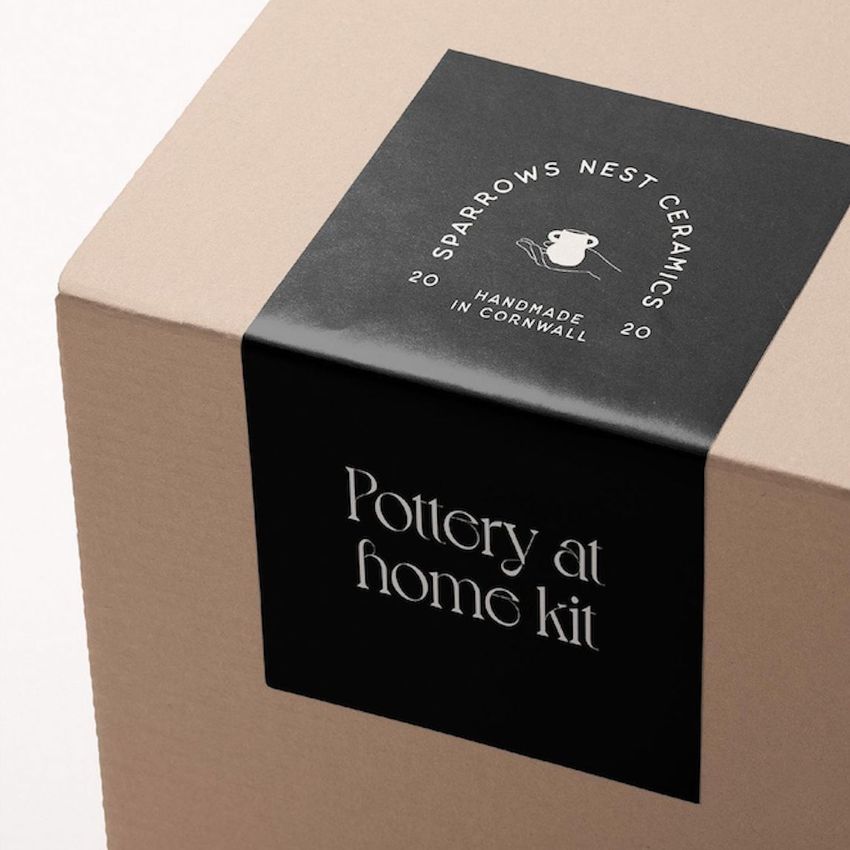 Minimalist cardboard box mockup with a black sticker displaying white decorative text and a pottery icon on a plain background.