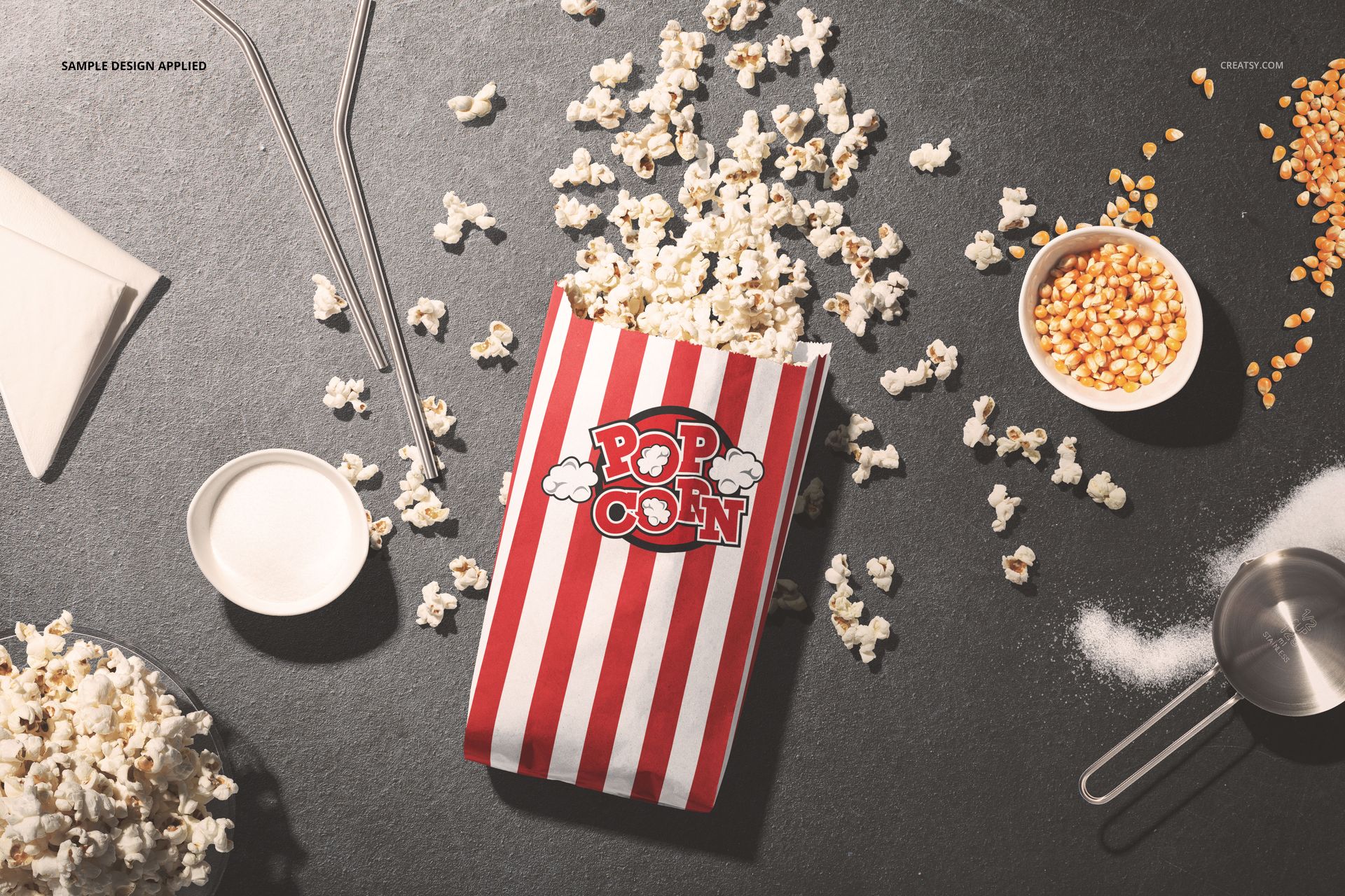 Red and white striped popcorn paper bag mockup on a dark surface, surrounded by popcorn kernels and utensils.