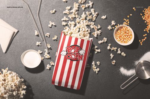 Red and white striped popcorn paper bag mockup on a dark surface, surrounded by popcorn kernels and utensils.